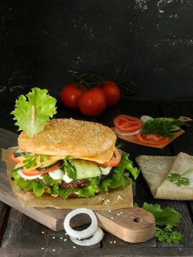 sandwich with ham and vegetables. Burger with sesame bun, cucumber, tomato, cheese, onion, sauce and salad, lies on a dark cutting board on a black background. Photo for the recipe.Tomatoes lie behind