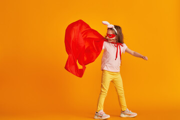 fun little girl is playing in superhero costume. baby is dressed in rabbit ears, red cape, mask, holding out her hand on yellow background. concept girl's superpowers, feminism, striving for victory