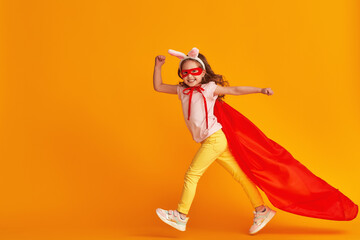 fun little girl is playing superhero game. baby is dressed in rabbit ears, red cape and mask, reaches out and runs on yellow background. concept girl's superpowers, feminism, striving for victory