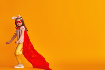fun little girl is playing superhero game. baby is dressed in rabbit ears, red cape and mask, reaches out and runs on yellow background. concept girl's superpowers, feminism, striving for victory