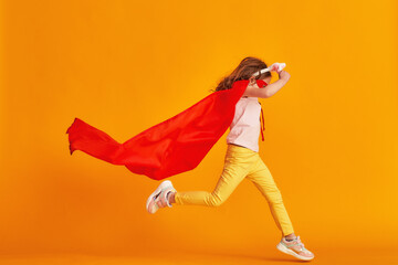 fun little girl is playing superhero game. baby is dressed in rabbit ears, red cape and mask, reaches out and runs on yellow background. concept girl's superpowers, feminism, striving for victory
