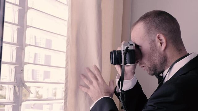 Man in tuxedo taking photos from a window. Handsome guy in black suit holding a camera and photographing people from a window with shutters. undercover agent hiding behind a curtain and spying.