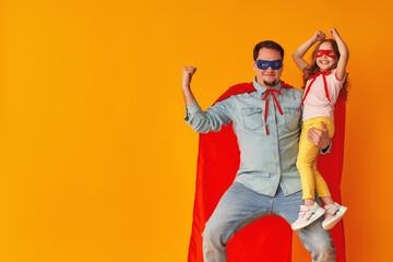 Fototapeta na wymiar serious father with a beard, a superhero holding a little baby girl, together dressed in superman costumes, preparing for a children's party, isolated on a yellow wall. Strong heroes. copy-space