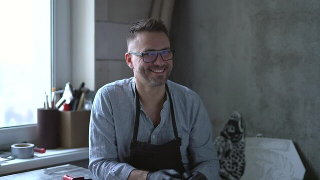 Portrait of bearded middle aged male lithograph artist sitting on a background of workplace. Linocut master in apron smiling and looking straight in camera. 4K footage