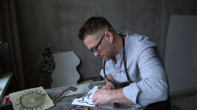 Linocut artist printmaking handmade work. Bearded middle aged man in apron making linocut using cutting instrument. 4k footage