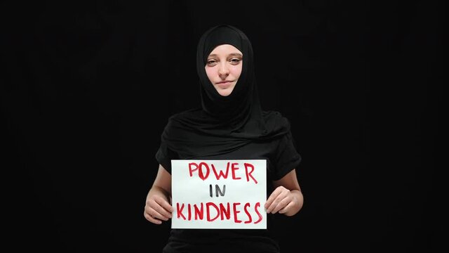 Portrait of smiling Muslim woman in hijab holding Power in kindness sign looking at camera. Positive confident young lady posing at black background. Gender equality and human rights concept.