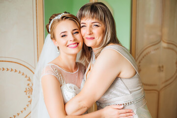 A beautiful bride in a wedding dress poses with her mother in the morning at home before the ceremony.