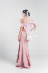 Fototapeta na wymiar full length view of bride in pink dress and veil posing on grey