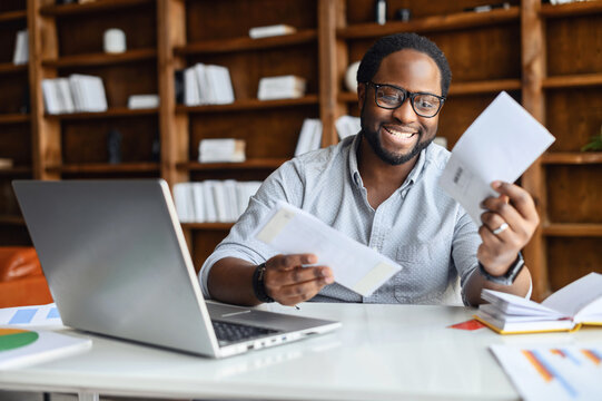 Smiling black man in the office in anticipation of the long-awaited letter about work, from relatives, received mailing envelopes, sitting at desk with laptop and documents with graphs, received news