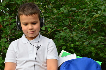 A schoolboy boy in a white shirt next to a briefcase in the park is playing music on headphones. Student learning listening audio tutorials in a park sitting on the grass in a park