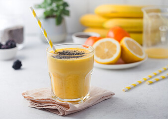 A yellow smoothie in a glass with a straw and chia seeds, fresh fruit in the background. Healthy...