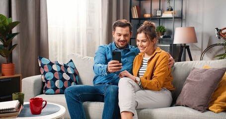 Obraz premium Lovely Caucasian young cute family couple wife and husband browsing online on smartphone resting at home sitting on sofa in modern living room, chatting and smiling spending time together love concept