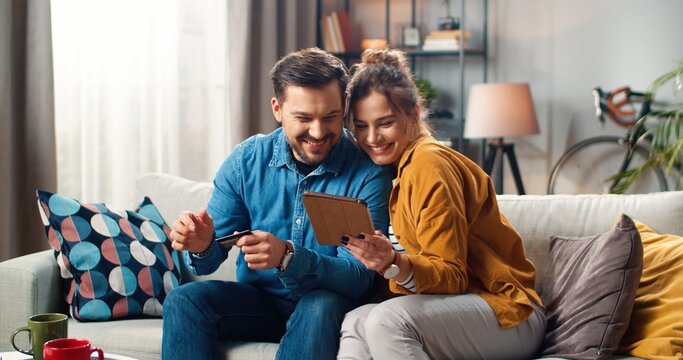 Cheerful Happy Young Caucasian Married Couple Man And Woman Buying Online Typing On Tablet Paying With Credit Card While Sitting On Couch In Cozy Room At Home, Satisfied Buyers, Internet Shopping