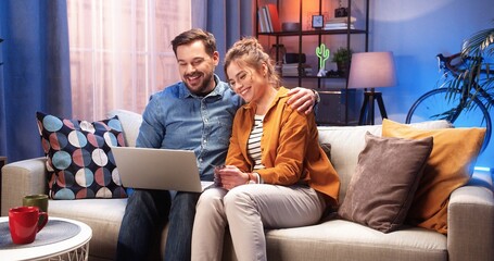 Happy young Caucasian family couple handsome man and beautiful woman talking sitting on sofa in living room at home in evening surfing internet on laptop choosing something. Family leisure concept