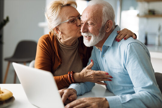 Mature woman kissing her mature husband while he uses a computer at home