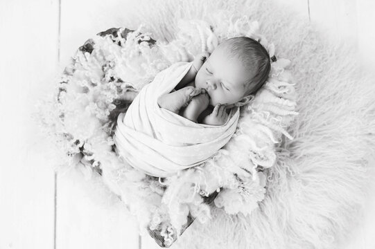 Newborn baby girl wrapped in wrap with flower pattern on wool fluffy blanket in wooden bowl in the shape of heart on white wood floor. Sweet infant sleeping in props for newborn photography. Black and