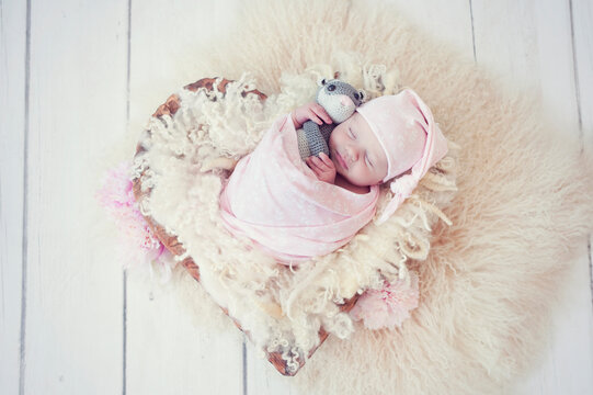 Newborn baby girl wrapped in pink wrap on wool fluffy blanket in wooden bowl in the shape of heart on white wood floor. Sweet infant sleeping in props for newborn photography. Baby with little sweet f