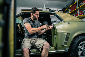 Young man removing interior trim panels from an old vintage car from the 60s or 70s in his home garage. Tools are seen around.