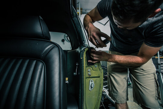 Young Man Removing Interior Trim Panels From An Old Vintage Car From The 60s Or 70s In His Home Garage. Tools Are Seen Around.