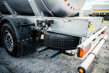 Truck with trailer, tank with flammable liquid, sunny day outside, metallic color container, blue sky with white clouds, gravel
