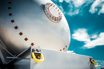 Truck with trailer, tank with flammable liquid, sunny day outside, metallic color container, blue sky with white clouds, gravel