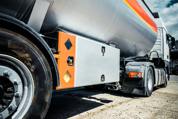 Truck with trailer, tank with flammable liquid, sunny day outside, metallic color container, blue sky with white clouds, gravel