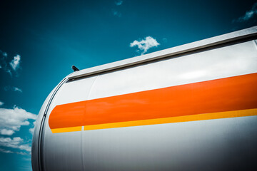 Truck with trailer, tank with flammable liquid, sunny day outside, metallic color container, blue sky with white clouds, gravel