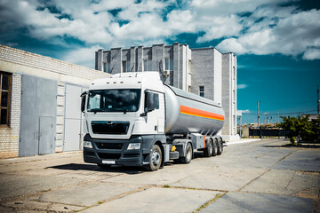 Truck with trailer, tank with flammable liquid, sunny day outside, metallic color container, blue sky with white clouds, gravel