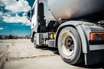 Truck with trailer, tank with flammable liquid, sunny day outside, metallic color container, blue sky with white clouds, gravel