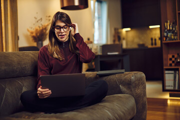 Cute pregnant brunette sitting on the sofa at her home with laptop in the lap and putting on earphones.