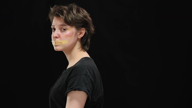 Young abused woman with bruises on face turning to camera with Stop violence sign on mouth. Portrait of desperate helpless Caucasian victim of domestic violence at black background on the left.