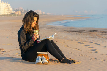 Joven mujer leyendo, tomando café y observando el amanecer junto a la orilla del mar.