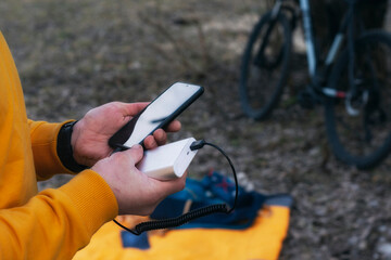 A tourist charges a smartphone with a power bank on the background of a backpack and a bicycle in nature.