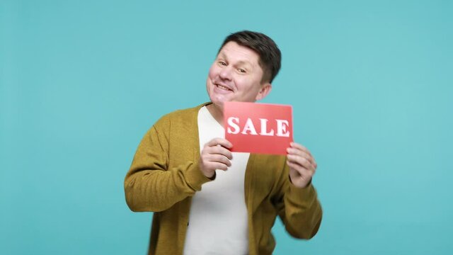 Delighted joyful mature man in white t-shirt and cardigan dancing holding in hands red template with sale inscription, happy satisfied with discounts. Indoor studio shot isolated on blue background
