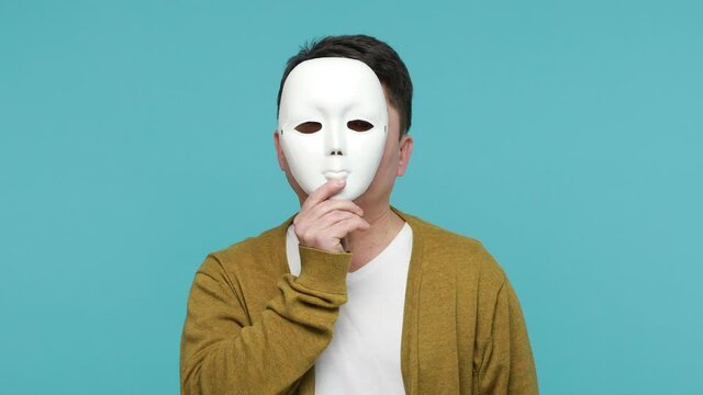 Middle aged dark haired man in white t-shirt and cardigan putting off mask showing different emotions, duplicity, hypocrisy. Indoor studio shot isolated on blue background