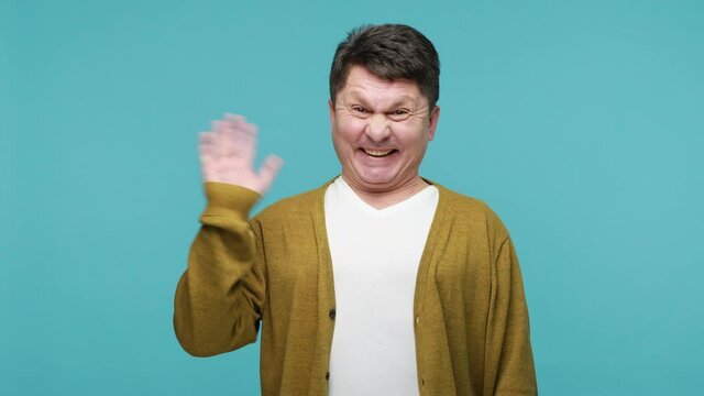 Extremely cheerful hospitable middle aged man waving hand saying hi, glad to see you, looking at camera with friendly expression and smiling. Indoor studio shot isolated on blue background