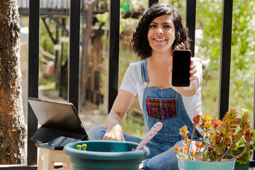 Young Hispanic woman on her terrace showing a mobile phone with a blank screen-planter using...