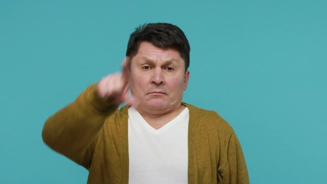 Unlucky annoyed mature dark haired man in t-shirt and cardigan looking sadly and showing loser gesture on his forehead, experiencing life difficulties. Indoor studio shot isolated on blue background