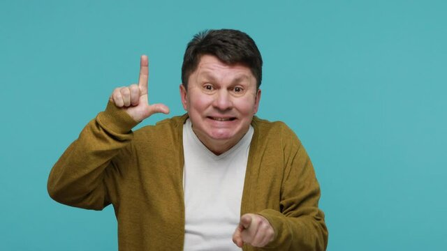 Middle aged man abuser showing loser gesture and pointing on you, blaming accusing for unsuccess, expressing disrespect, mocking your failure. Indoor studio shot isolated on blue background