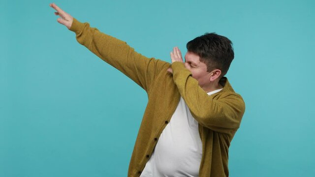 Positive mature brunet man in white t-shirt and cardigan having fun dancing and moving to rhythm, dabbing raising hands, making dubdance gesture. Indoor studio shot isolated on blue background