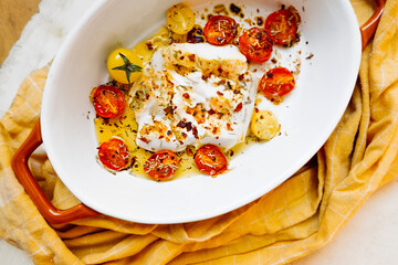 close-up baked molten feta cheese with small tomatoes in olive oil with herbs and spices in ceramic orange baking dish and yellow napkin. simple cheese dishes. vegan menu, selective focus