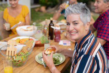 Portrait of smiling caucasian senior woman holding hotdog at table with family having meal in garden