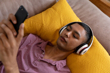 Obraz premium Mixed race man lying on couch wearing headphones and using smartphone