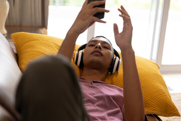 Mixed race man lying on couch wearing headphones and using smartphone