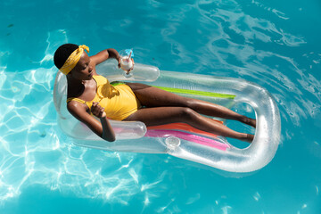 African american woman in swimming pool sunbathing on inflatable and holding drink
