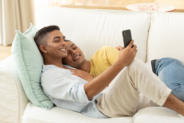 Diverse gay male couple sitting on sofa using smartphone and embracing