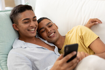 Diverse gay male couple sitting on sofa using smartphone and embracing