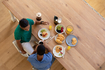 Diverse gay male couple eating breakfast together and talking