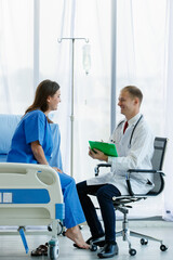 Woman caucasian patient sitting on bed and caucasian doctor sitting on chair are talking, both are smiling look happy. Doctor man carrying stethoscope on neck and hold writing on note in office room
