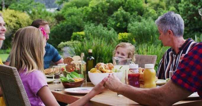 Smiling caucasian family holding hands saying grace before celebration meal together in garden